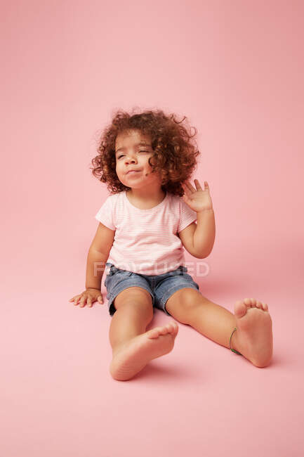 Cute thoughtful toddler girl with curly hair in casual clothe looking away while sitting on pink background — Stock Photo