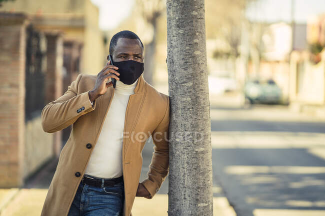 Irreconocible reflexivo joven negro en traje de moda y máscara de la cara apoyada en el árbol en la calle de la ciudad y mirando hacia otro lado mientras tiene conversación telefónica - foto de stock