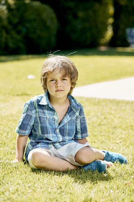 Criança séria em camisa quadriculada olhando para a câmera enquanto sentada no gramado — Fotografia de Stock