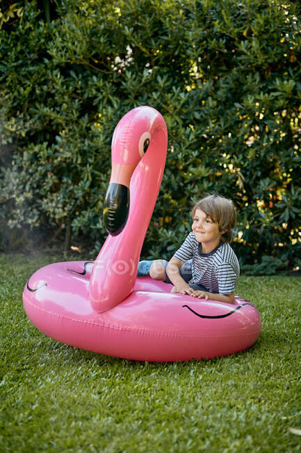 Повне тіло маленького хлопчика в повсякденному одязі лежить на надувному рожевому фламінго, розважаючись на трав'янистому газоні в парку — стокове фото