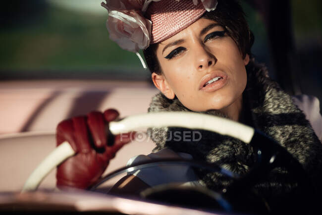 Crop trendy female with flower on hat and leather gloves touching steering wheel in automobile while looking at camera — Stock Photo