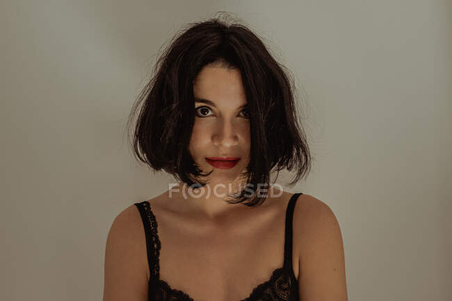 Heitere Frau im schwarzen Spitzenbody sitzt auf Hocker im Raum vor weißer Wand und blickt in die Kamera — Stockfoto
