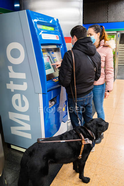 Полное тело женщины в маске помогает мужчине с слепотой стоять с собакой-поводырем в покупке билета на метро в торговом автомате — стоковое фото