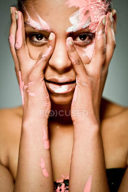 Modello femminile etnico creativo con viso imbrattato di vernice rosa e bianca che tocca le guance e guarda la fotocamera su sfondo grigio in studio — Foto stock