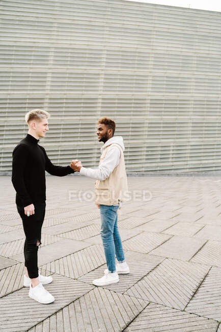 Side view of stylish young diverse male friends in trendy outfits standing on paved street in city and shaking hands — Stock Photo