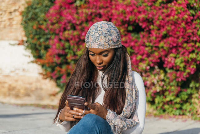 Mujer negra en traje casual sentada en el parque urbano y charlando en las redes sociales a través de un teléfono inteligente - foto de stock