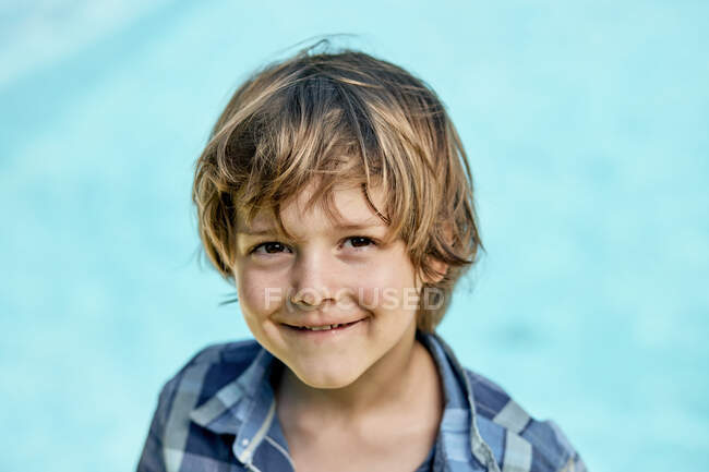 Adorable niño pequeño con el pelo rubio con elegante camisa a cuadros sonriendo y mirando a la cámara mientras está de pie sobre el fondo azul a la luz del sol - foto de stock