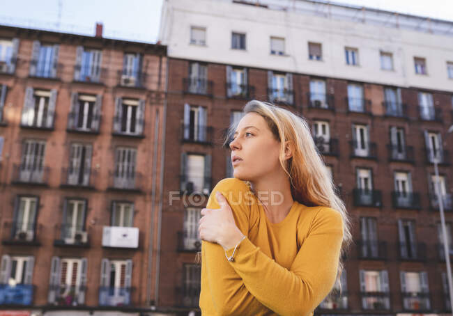 Baixo ângulo de jovem sonhadora com longos cabelos loiros em roupas casuais abraçando a si mesma e olhando para longe enquanto estava na rua da cidade perto do edifício de tijolos — Fotografia de Stock