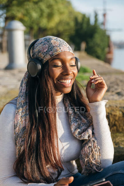 Entzückte Afroamerikanerin sitzt im Park und hört Musik über Kopfhörer, während sie Lieder mit geschlossenen Augen genießt — Stockfoto