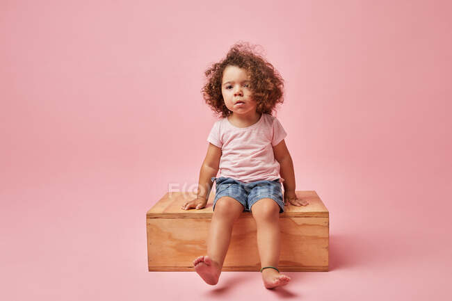 Charmantes Barfußkind in T-Shirt und Jeanshose mit lockigem Haar, das auf einer Holzplattform sitzt und wegschaut — Stockfoto