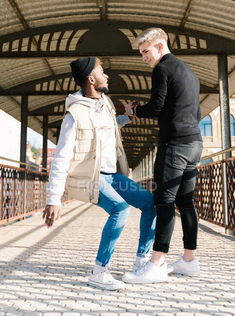 Side view of young stylish multiracial male friends in trendy outfits shaking hands and having fun while standing together on paved walkway — Stock Photo
