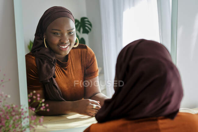 Glückliche junge, stylische Afrikanerin im traditionellen Hijab, die sich auf einen Kosmetiktisch lehnt und zu Hause im Spiegel im Sonnenlicht sieht — Stockfoto
