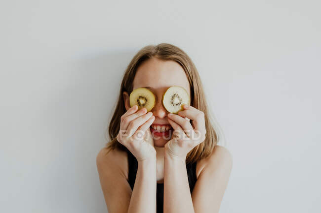 Fröhliches Mädchen in lässiger Kleidung lächelt, während sie die Augen mit Kiwi-Scheiben vor weißem Hintergrund bedeckt — Stockfoto