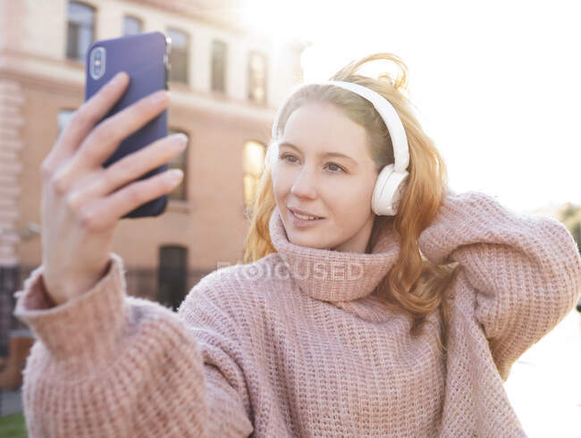 Уверенная молодая женщина в теплых свитерах и наушниках трогает длинные волнистые волосы, делая селфи на смартфоне в солнечный день в городе — стоковое фото