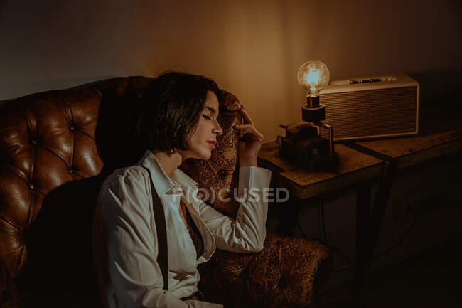Vue latérale d'une femme sereine relaxante dans un vieux fauteuil en cuir dans une pièce vintage avec ampoule lumineuse et yeux fermés — Photo de stock