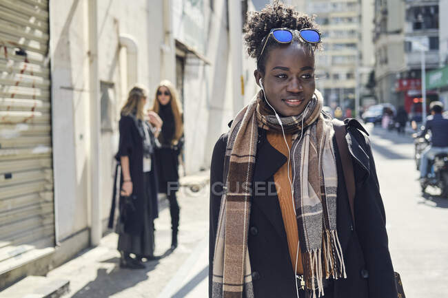 Jovem afro-americana em roupas elegantes e óculos de sol olhando para longe na estrada da cidade contra amigas irreconhecíveis no fundo — Fotografia de Stock