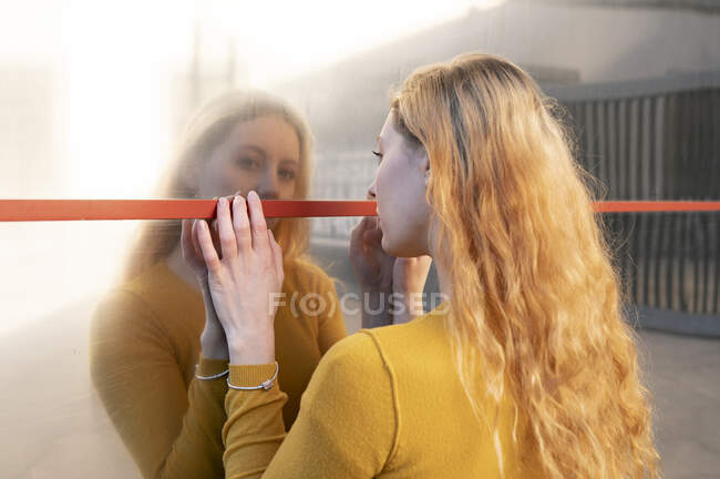 Vista trasera de la joven con el pelo ondulado ling en ropa casual de pie en la calle de la ciudad cerca del viejo espejo y mirando a la cámara - foto de stock