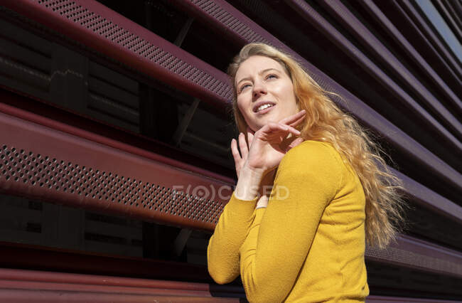 Vista lateral de la joven milenaria pensativa en ropa casual con el pelo ondulado largo y rubio de pie cerca de la valla de metal rojo - foto de stock