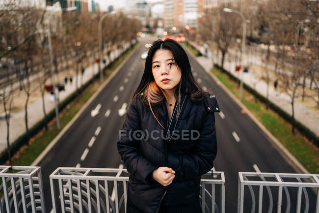 Cabello largo morena asiática mujer de pie en un puente y mirando a la cámara - foto de stock
