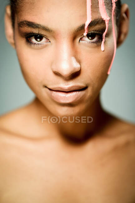 Creativo modello etnico femminile con vernice rosa gocciolante sul viso guardando la fotocamera su sfondo grigio in studio — Foto stock
