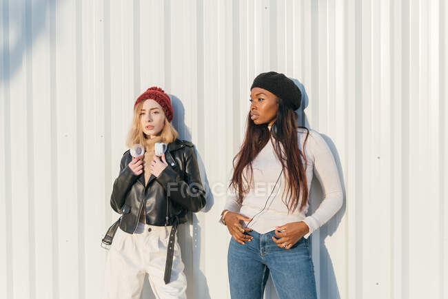 Cool multiracial female models wearing trendy clothes standing near metal wall in city on sunny day — Stock Photo