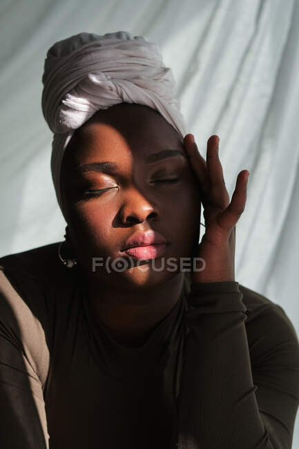 Graciosa jovem mulher africana com excesso de peso no tradicional turbante tocando o rosto com os olhos fechados na luz solar — Fotografia de Stock