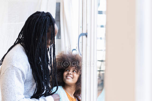 Vista lateral del hombre afroamericano de pie detrás de la chica de pelo rizado cerca de la ventana y mirándose entre sí - foto de stock