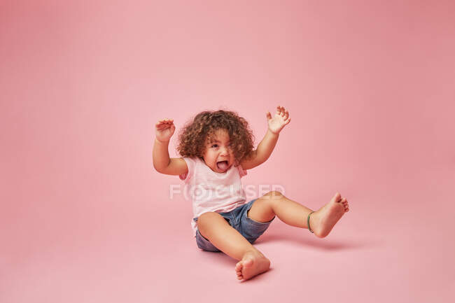 Mignon joyeux bambin fille avec des cheveux bouclés dans des vêtements décontractés avoir du plaisir à faire des visages tout en étant assis sur le sol regardant loin sur fond rose — Photo de stock