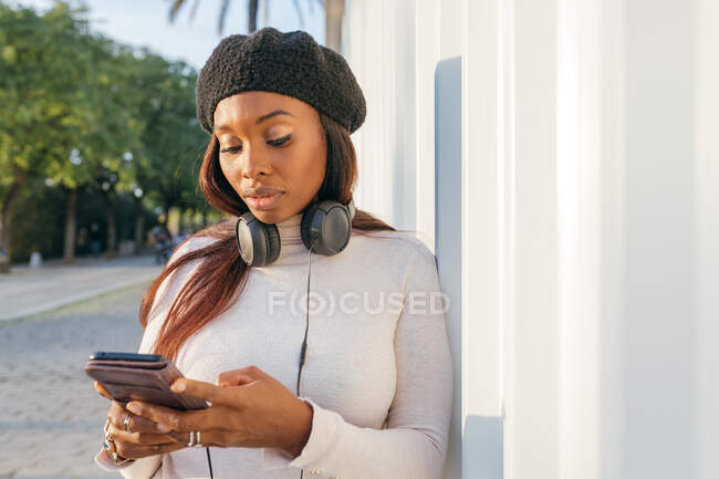 Femme noire détendue avec écouteurs sur le cou s'appuyant sur la construction et la navigation téléphone mobile sur la rue de la ville — Photo de stock