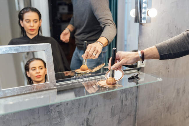 Crop visagiste masculino anónimo usando polvo y cepillo mientras hace maquillaje para cliente joven concentrado sentado frente al espejo en el salón de belleza moderno - foto de stock