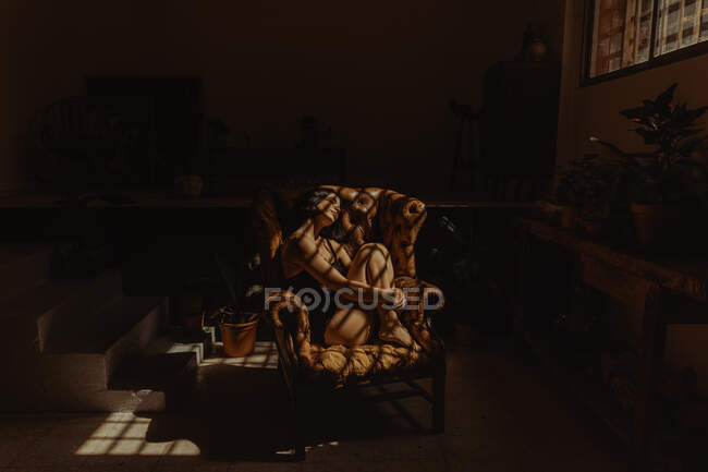 Side view of peaceful female in nightgown sitting in leather armchair lit by sunlight and relaxing with closed eyes — Stock Photo
