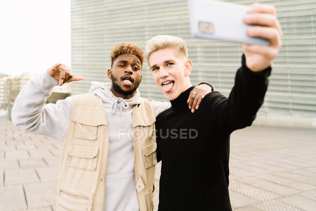 Glücklicher junger afroamerikanischer Typ in trendiger Kleidung, der lächelt, während er auf der Straße ein Selbstporträt mit der Hand auf der Schulter eines männlichen Freundes macht und die Zunge herausstreckt — Stockfoto