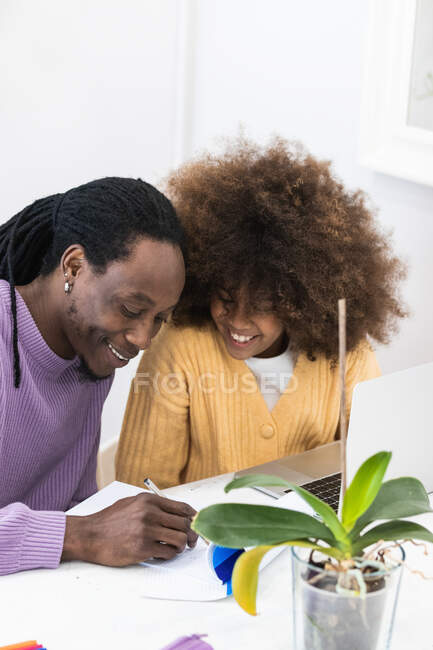 Sonriente papá afroamericano sentado en la mesa con la niña surfeando netbook mientras se prepara la tarea para la escuela - foto de stock