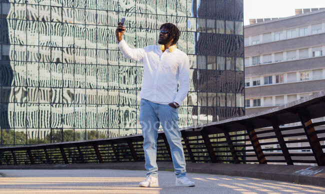 Masculine African American male in stylish outfit with hand in pocket taking self portrait on cellphone against urban buildings — Stock Photo