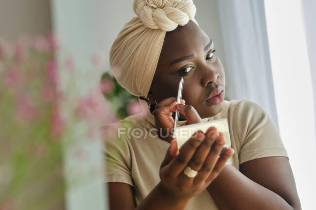 Concentrated young African lady in traditional turban holding small mirror in hand while applying eyeliner in light bedroom — Stock Photo