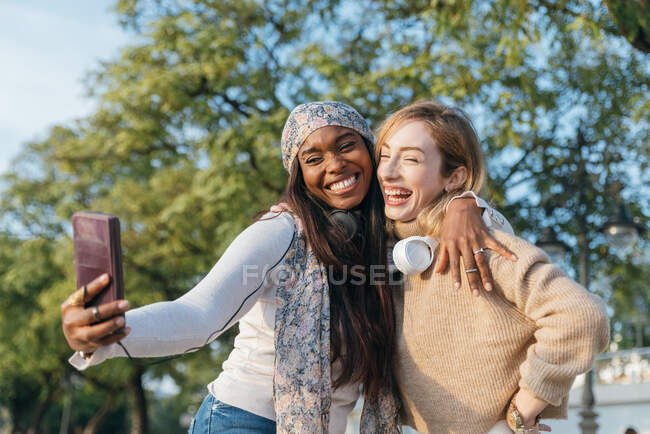 Mulher tomando selfie de amigo feminino branco enquanto relaxa no parque na cidade — Fotografia de Stock