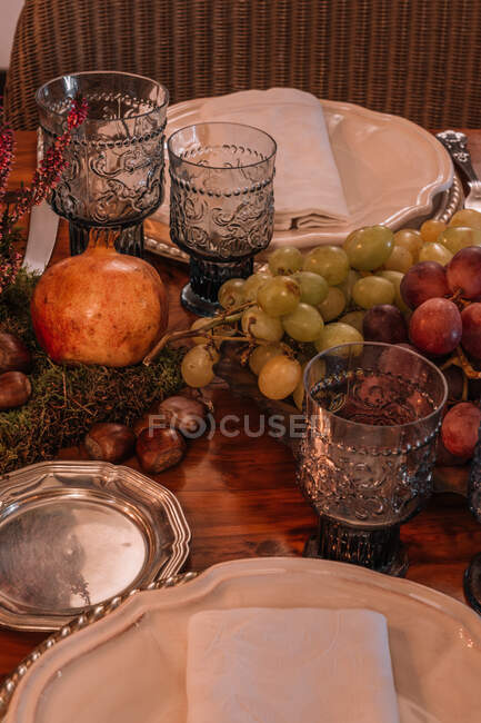 Grand angle de verres en cristal près de la plaque et des couverts placés sur la table décorée de raisins Calluna vulgaris fleurs et grenade — Photo de stock