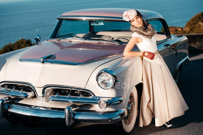 Cuerpo completo de mujer elegante con guantes de cuero vestido largo y sombrero de pie cerca de coche vintage contra el mar - foto de stock