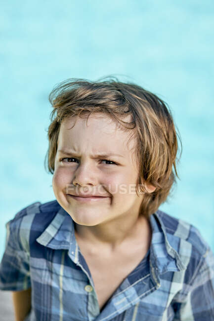 Funny little boy with blond hair in checkered shirt frowning and looking at camera against blue background — Stock Photo