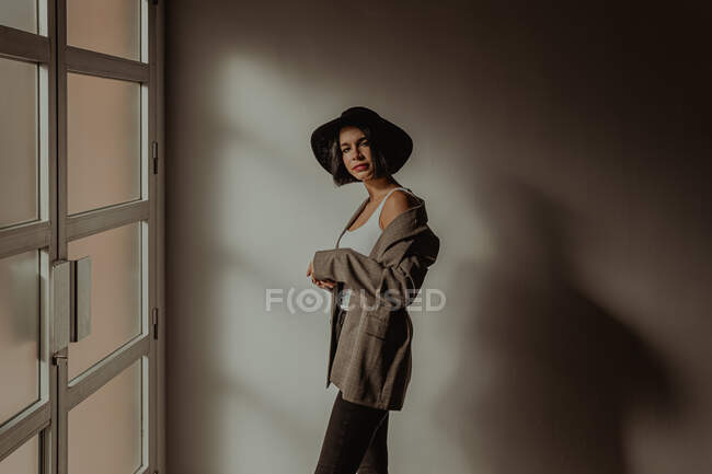 Vista lateral de determinada fêmea na roupa da moda em pé na sala perto da janela e olhando para a câmera — Fotografia de Stock