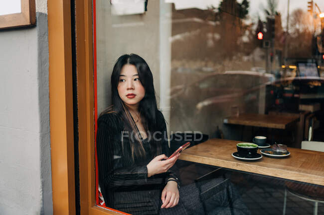 Langhaarige brünette asiatische Frau, die einen Kaffee in einem Coffeeshop hat, während sie ein Handy sucht — Stockfoto