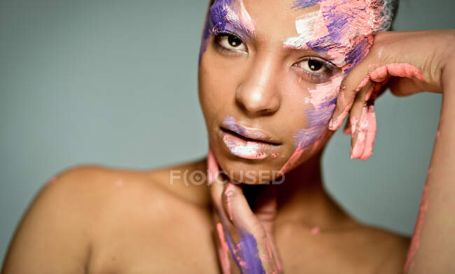 Creative ethnic female model with face smeared with pink and white paint leaning on hand and looking at camera on gray background in studio — Stock Photo