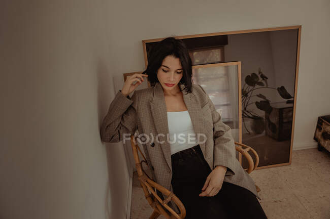 Удумлива жінка в модному одязі, сидячи на стільці, торкаючись волосся і дивлячись в роздуми — стокове фото