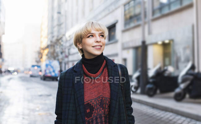 Attraente donna positiva in abbigliamento alla moda e cappotto caldo in piedi con le mani in tasca sulla strada innevata della città e guardando altrove nella chiara giornata invernale a Madrid, Spagna — Foto stock