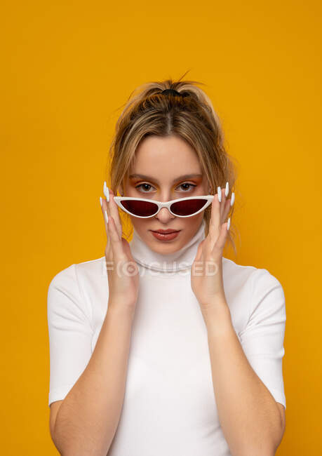 Magnifique jeune femme en vêtements blancs abaissant les lunettes de soleil à la mode et regardant la caméra tout en se tenant debout sur fond jaune en studio — Photo de stock