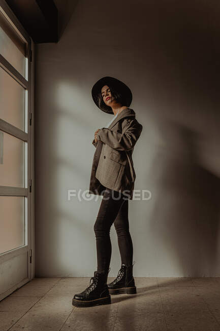Side view low angle of determined female in trendy outfit standing in room near window and looking at camera — Stock Photo