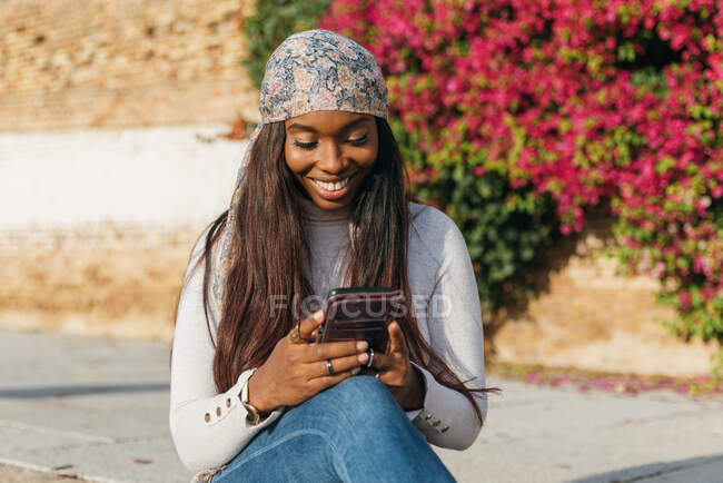 Чорна жінка в повсякденному вбранні сидить у міському парку і спілкується в соціальних мережах через смартфон — стокове фото