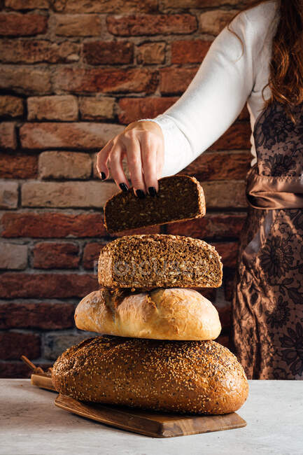 Panadero femenino anónimo de cosecha que muestra pan fresco suave con semillas crujientes en la mesa en la panadería - foto de stock