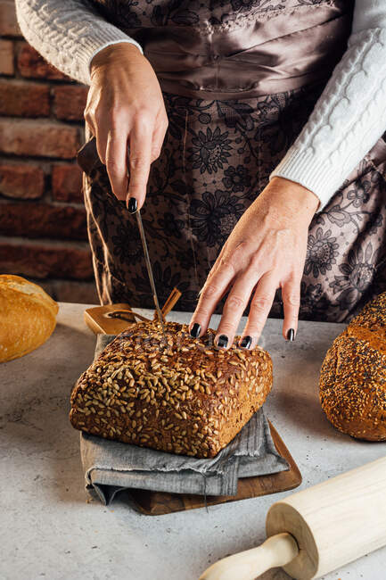 Crop anonymous female baker with knife cutting loaf of fresh bread with sunflower seeds on table in bakehouse — Stock Photo