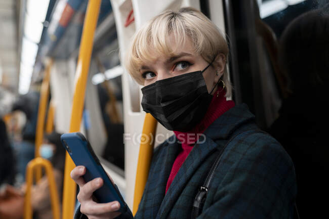 Calm young female with short hair in warm jacket and protective mask browsing mobile phone and looking away while riding metro train in Madrid, Spain — Stock Photo